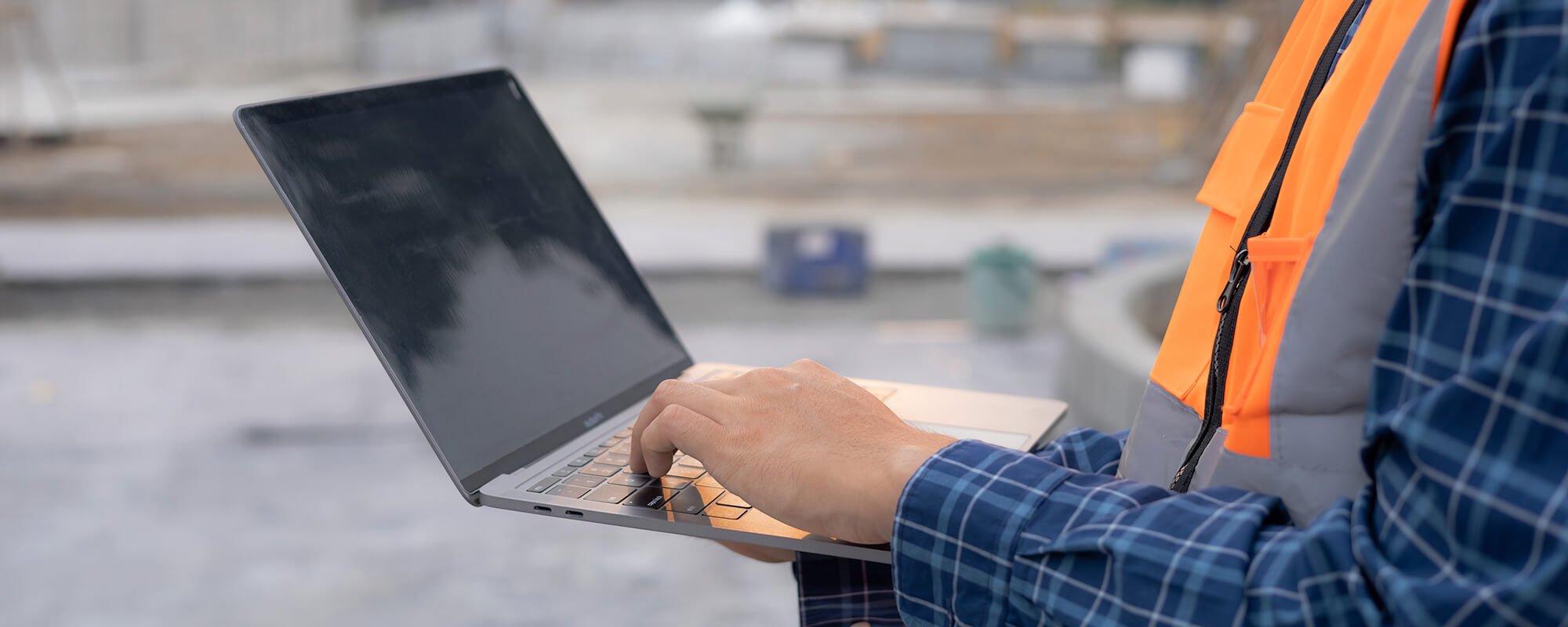 L'image montre une personne travaillant sur un ordinateur portable sur ce qui semble être un chantier de construction. La personne porte une chemise à carreaux et un gilet de sécurité orange, ce qui indique qu'elle est probablement un travailleur de la construction ou un superviseur. L'ordinateur portable est ouvert et semble être utilisé activement, ce qui suggère que la personne pourrait être en train de consulter des plans, de gérer des documents ou de communiquer avec d'autres membres de l'équipe. L'arrière-plan est flou, mais on peut distinguer des matériaux de construction et un environnement de chantier, ce qui renforce l'idée que la personne est sur un site de travail actif.