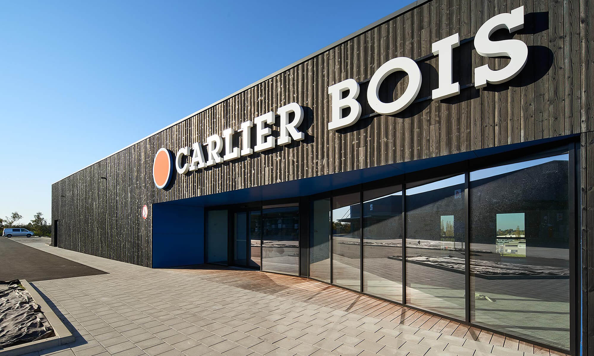 Cette image montre l'extérieur d'un bâtiment commercial avec une enseigne indiquant Carlier Bois. Le bâtiment a une façade moderne avec des panneaux verticaux en bois sombre et des fenêtres vitrées à l'avant. Le logo de l'entreprise, qui semble être un cercle avec un motif à l'intérieur, est affiché à côté du nom Carlier Bois. Le bâtiment est situé dans une zone qui semble être un parc industriel ou commercial, avec un parking et une route adjacente. Le ciel est dégagé et bleu, ce qui suggère une journée ensoleillée.